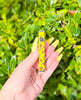 A hand holding a yellow 510 threaded battery with a ducky design, against a backdrop of green foliage.