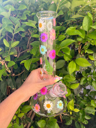 A hand holding a dried floral glass bong in front of a bush