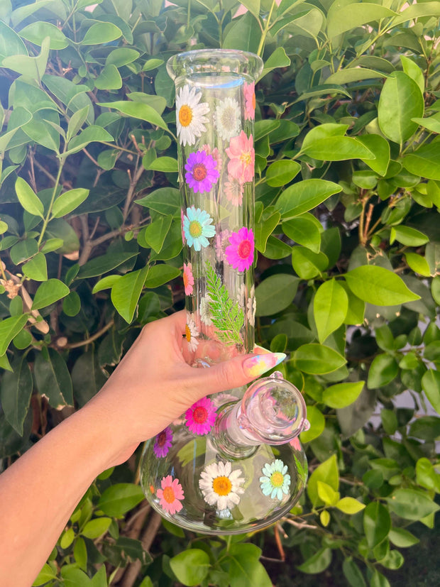 A hand holding a dried floral glass bong in front of a bush
